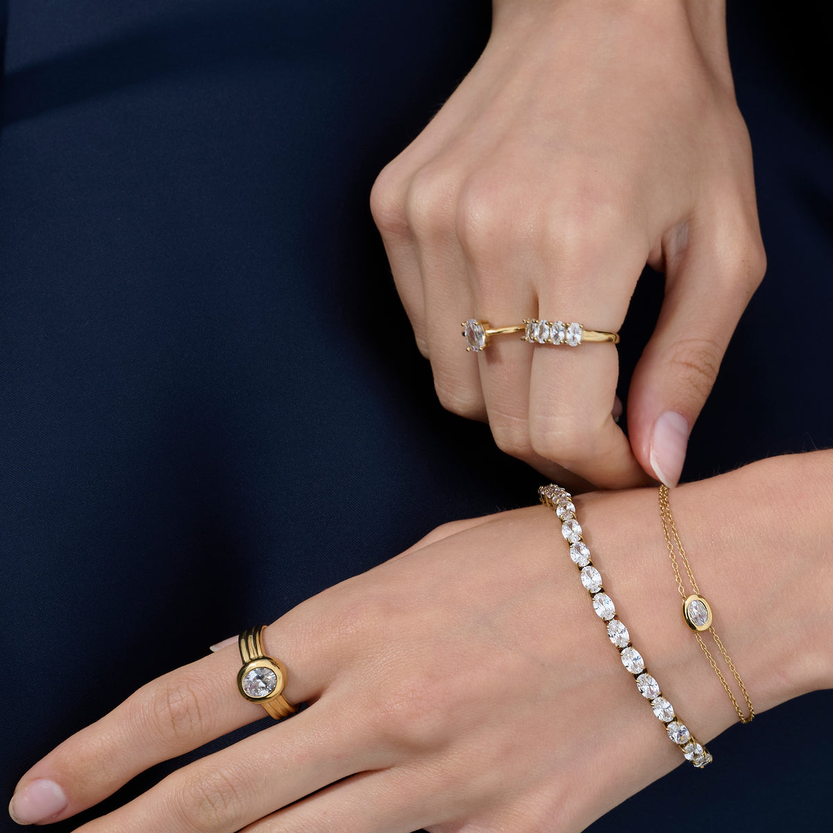 Close-up lifestyle shot of a woman's hands wearing a gold ring with a round clear stone and ridged band, paired with two stone-set gold rings and two gold bracelets against a navy background.
Photo lifestyle en gros plan des mains d'une femme portant une bague dorée sertie d'une pierre transparente ronde à anneau strié, agencée avec deux bagues dorées serties de pierres et deux bracelets dorés sur fond bleu marine.