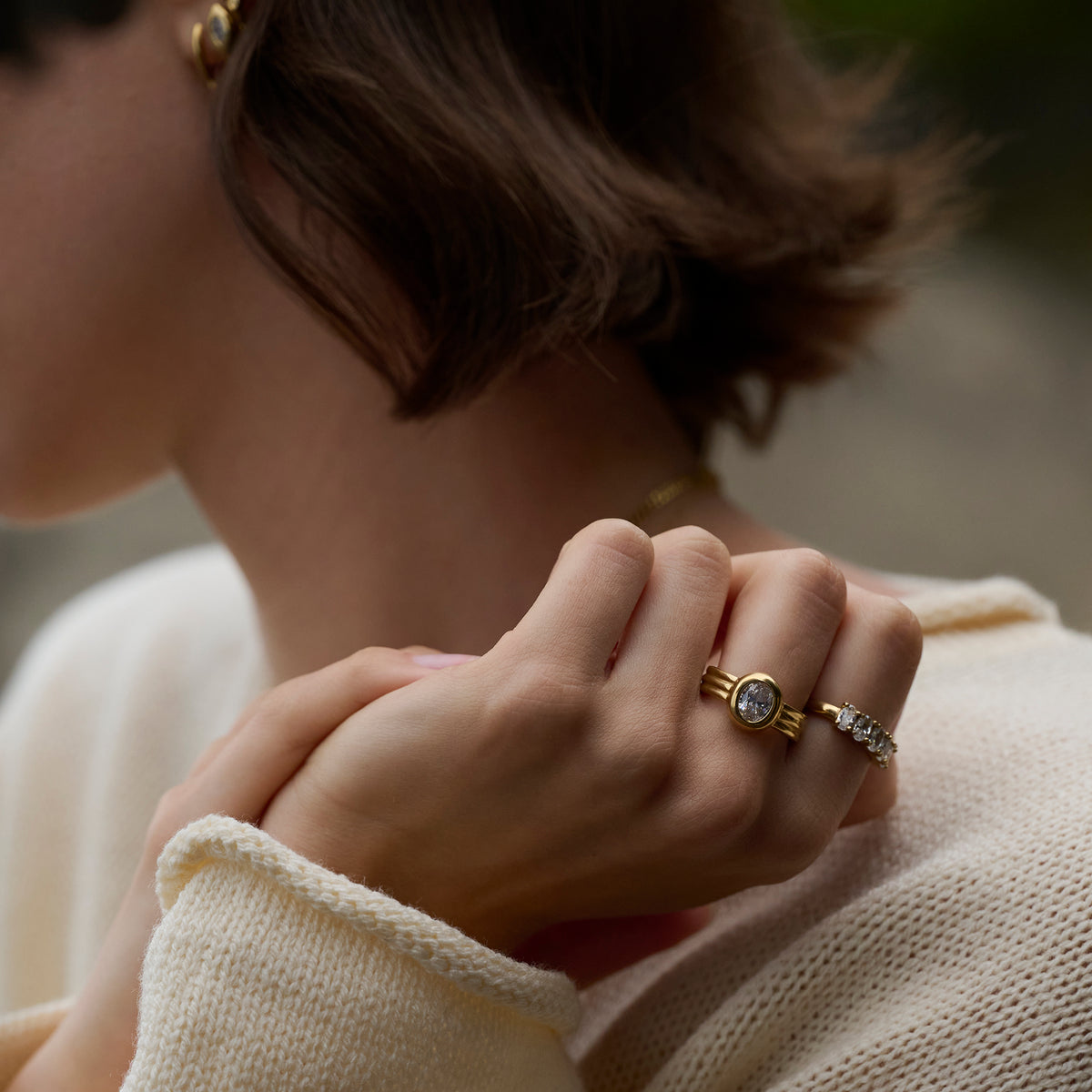 Close-up lifestyle shot of a woman's hands wearing a gold ring with a round clear stone and ridged band, paired with a stone-set gold ring and a cream knit sweater.
Photo lifestyle en gros plan des mains d'une femme portant une bague dorée sertie d'une pierre transparente ronde à anneau strié, agencée avec une bague dorée sertie de pierres et un tricot crème.