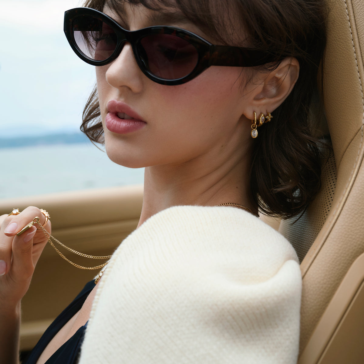 Close-up lifestyle shot of a woman seated in a car by the water, wearing layered gold earrings set with clear stones, a fine gold chain necklace and gold rings, styled with sunglasses and a cream knit.
Photo lifestyle en gros plan d'une femme assise dans une voiture au bord de l'eau, portant des boucles d'oreilles dorées superposées serties de pierres transparentes, un collier à chaîne fine dorée et des bagues dorées, agencés avec des lunettes de soleil et un tricot crème.